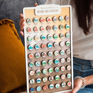 May include: A white board with the text "Mark and Marie's National Park Adventures" at the top. The board is filled with small, round, colorful wooden magnets, each representing a different national park in the United States.