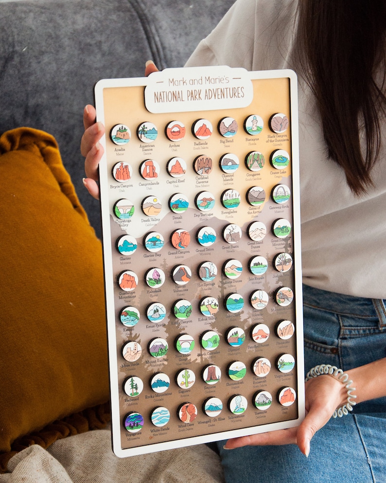 May include: A white board with the text "Mark and Marie's National Park Adventures" at the top. The board is filled with small, round, colorful wooden magnets, each representing a different national park in the United States.