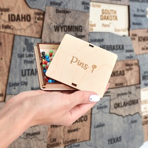 May include: A wooden box with a lid that says "Pins 9" on the top. The box is open and filled with colourful push pins. The box is being held by a hand in front of a wooden map of the United States.