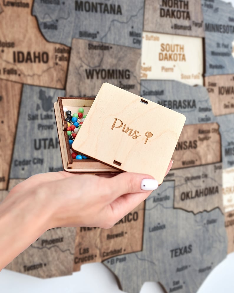 May include: A wooden box with a lid that says "Pins 9" on the top. The box is open and filled with colourful push pins. The box is being held by a hand in front of a wooden map of the United States.