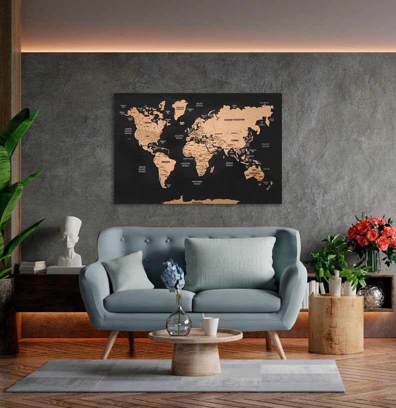 May include: A black and gold world map with a gold foil finish is hanging on a wall above a light blue sofa with two cushions. The sofa is in a living room setting with a wooden coffee table and a wooden side table.