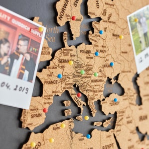 May include: A corkboard world map with push pins marking locations. The map is brown and has a dark gray background. There are two photos attached to the corkboard, one of a couple and one of a group of people.