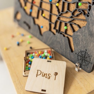 May include: A wooden map of the United States with a cork surface and a compass design. The map is decorated with colorful push pins. A small wooden box with the word "Pins" and a pin icon is next to the map.