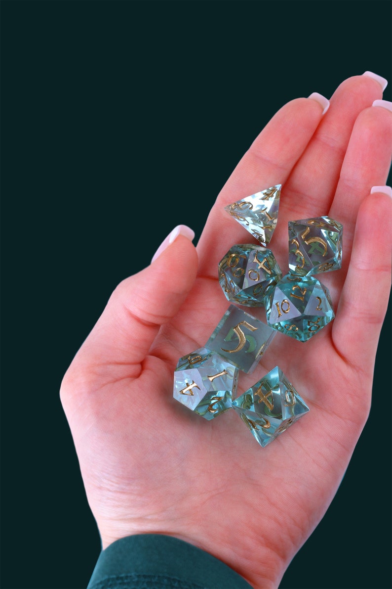 May include: A set of seven teal blue and gold dice with various numbers and symbols. The dice are made of clear glass with gold accents and are sitting in a hand.