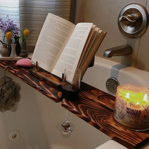 May include: A wooden bath tray with a book holder, a wine glass holder, and a candle holder. The tray is made of dark wood and has a natural finish. The book holder is made of metal and has a black finish. The wine glass holder is made of metal and has a silver finish. The candle holder is made of glass and has a clear finish. The candle is white and has a gold label that says "Winter Woods".