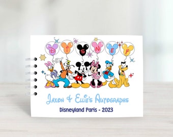 Puede incluir: Un libro de autógrafos blanco con una ilustración colorida de Mickey Mouse, Minnie Mouse, Donald Duck, Goofy, Pluto y Daisy Duck. El libro tiene una encuadernación en espiral y el texto "Jaxon & Ellie's Autographs Disneyland Paris - 2023" impreso en la portada.