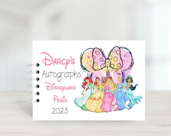 Puede incluir: Un libro de autógrafos blanco con un lazo rosa y amarillo y un dibujo de princesas Disney. El libro dice "Darcy's Autographs Disneyland Paris 2023".
