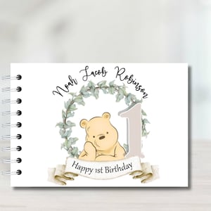 May include: A white spiral-bound book with a Winnie the Pooh design. The cover features a watercolor illustration of Pooh Bear, a number 1, and the text "Happy 1st Birthday." The names "Noah Jacob Robinson" are above the illustration.