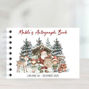 May include: A festive autograph book with a spiral binding. The cover displays a Christmas scene with Santa, elves, and a reindeer in a snowy setting. The text "Mable's Autograph Book" is in red script, with "LAPLAND UK - DECEMBER 2025" below.