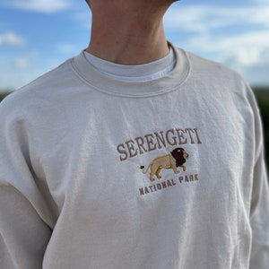 Op de afbeelding: Beige sweatshirt met geborduurde tekst "Serengeti National Park" en een leeuwenafbeelding. De crew neck sweatshirt heeft een neutrale kleur. De achtergrond toont een vage buitenscène.