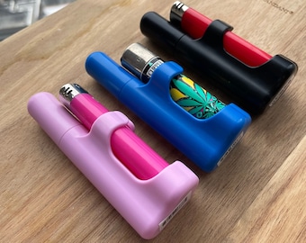 May include: Three different colored cases for lighters. The cases are made of plastic and have a clip on the side. The cases are pink, blue, and black. The black case has a red lighter inside. The blue case has a green and yellow patterned lighter inside. The pink case has a pink lighter inside.