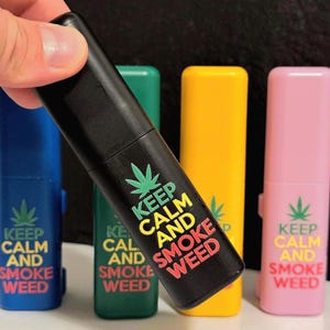 May include: A black, rectangular lighter with the text "KEEP CALM AND SMOKE WEED" in white, yellow, and red. The lighter is held in front of a blue, green, yellow, and pink lighter, each with the same text and a cannabis leaf.