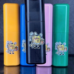 May include: A collection of colorful, rectangular containers in yellow, blue, black, pink, and green. Each container features a cartoon character sticker. The black container is held by a hand. The containers are arranged on a reflective surface.