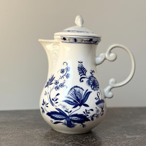 May include: A white ceramic coffee pot with a lid and handle, decorated with blue floral patterns. The pot has a curved spout and a decorative handle. The design features detailed blue flowers and foliage, creating a classic, elegant appearance.