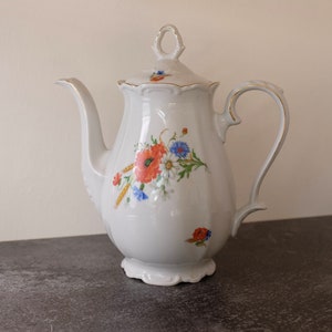 May include: White porcelain teapot with a floral design featuring red poppies, blue and white wildflowers, and wheat stalks. The teapot has a gold-trimmed handle and lid.