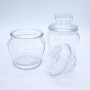 Puede incluir: Frascos de vidrio transparente con tapas. Un frasco está abierto, revelando un sello de goma blanco. El otro frasco tiene su tapa puesta. Los frascos están sobre un fondo blanco. Los frascos son probablemente para almacenamiento.