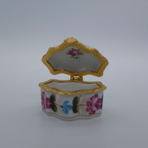 Puede incluir: Una pequeña caja de porcelana blanca con tapa dorada. La caja está decorada con diseños florales pintados a mano en tonos rosa, azul y verde.