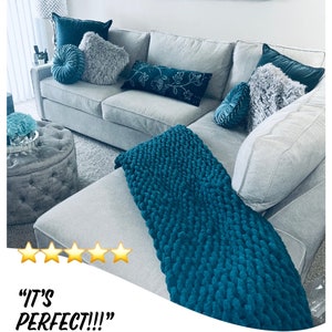 K&ouml;nnte beinhalten: Ein graues Ecksofa mit einer tealfarbenen gestrickten Wolldecke. Das Sofa hat mehrere Kissen in verschiedenen Farben und Texturen, darunter grau, teal und wei&szlig;. Das Sofa steht in einem Wohnzimmer mit einem kleinen Hocker und einer Wand mit Kunstwerken.