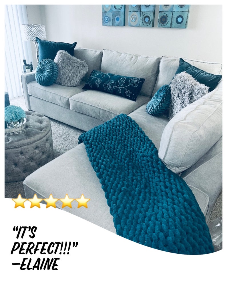 K&ouml;nnte beinhalten: Ein graues Ecksofa mit einer tealfarbenen gestrickten Wolldecke. Das Sofa hat mehrere Kissen in verschiedenen Farben und Texturen, darunter grau, teal und wei&szlig;. Das Sofa steht in einem Wohnzimmer mit einem kleinen Hocker und einer Wand mit Kunstwerken.
