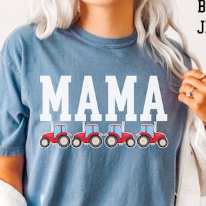 May include: Blue jean-colored t-shirt with the word "MAMA" in large white letters. Below the word "MAMA" are four red tractors with blue cabs. The text "BLUE JEAN" is in the upper right corner.