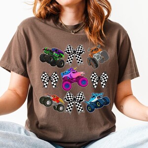 May include: A brown t-shirt featuring colorful monster truck graphics and checkered bow accents. The trucks are in various colors, including green, orange, pink, and blue. The shirt is worn by a person.