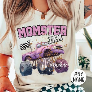 model is wearing a natural tshirt that says "Momster Jam" with a customized name signature "Mama" . Momster is in Pink Varsity Letters in a arch, Jam is in white letters, there is a Pink Monster Truck with a black and white checkered racing flag in the back of the truck. "mama" is in a handwritten text style. Family Matching sizes perfect for all the girls in your group or family. Birthday gift for her, Mother's Day Gift for Her.