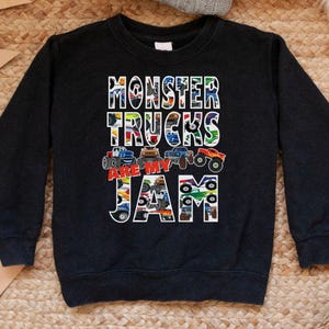 Op de afbeelding: Zwart sweatshirt met de tekst "MONSTER TRUCKS ARE MY JAM" in kleurrijke blokletters. De letters zijn gevuld met afbeeldingen van monstertrucks. Bovenop ligt een grijze gebreide muts.