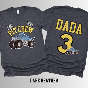 Two dark heather gray t-shirts. The front of one shirt has a blue shark monster truck with the varsity text 'Pit Crew' above it and 2 checkered flags. The back of the other shirt has the college varsity text 'Dada' in yellow with the number '3' below it with a small version of the shark truck.