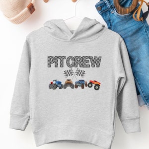 Op de afbeelding: Grijze hoodie voor peuters met een zwart-wit geblokt vlagontwerp en de tekst "PIT CREW". De hoodie heeft een kleine afbeelding van vier monstertrucks met geblokte vlaggen.