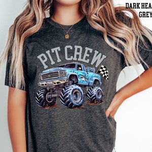 Pictured is a woman wearing a dark gray charcoal t-shirt which says "PIT CREW" in college varsity letters in light gray color. There is a blue monster truck with a shark on the side with a checkered flag. Suitable for parent of a birthday party child.
