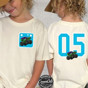 May include: Cream-colored t-shirt with a monster truck graphic on the front pocket and back. The back features the number "05" in blue. The front pocket graphic is a green and purple monster truck on a blue square with white stars. The shirt has the text "NATURAL".