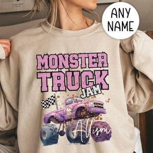 Op de afbeelding: Een beige sweatshirt met een roze en paarse monstertruck-afbeelding. De truck heeft een geblokte vlag, een eenhoorn en de woorden "Monster Truck Jam". De naam "Alison" staat in wit handschrift onder de truck.