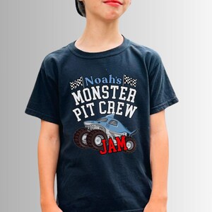Pictured is a boy wearing a navy blue t-shirt with the text "Noah's Monster Pit Crew JAM" and a graphic of a shark-shaped monster truck. The shirt has a crew neck and short sleeves. The child is wearing a black baseball cap.