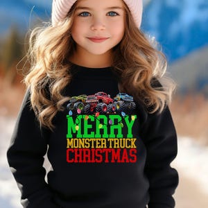 Op de afbeelding: Een zwarte sweatshirt met een afbeelding van drie groene, rode en blauwe monstertrucks, met de tekst "MERRY MONSTER TRUCK CHRISTMAS" in groen en rood. De sweatshirt wordt gedragen door een jong persoon met een roze muts.