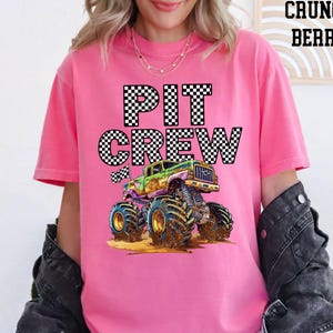 Lady is wearing a pink comfort colors t-shirt with a black and white checkered "PIT CREW" graphic and a green and purple cartoon monster truck with huge tires.