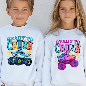 boy and girl are wearing matching white sweatshirts. The boy's says "ready to Crush" in Varsity Style multiple colors in an arch . underneath is a blue shark monster truck megalodon cartoon style. The girl's says Ready to Crush in Pink Varsity Letters - Multi-colored with a Pink Unicorn Monster Truck Sparkle Crash Smash cartoon style. Perfect to wear for car shows and truck racing events. Monster Truck Jam, Birthday Gifts and Pit Crew Parties. Parent and Sibling , grandparent, Family Matching