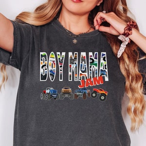 Pictured is a woman wearing a dark gray crop top which says BOY MAMA in large graphic letters filled with images of monster trucks on a white background and outlined in white. JAM is in red outlined in white and on the bottom are 4 multi-colored monster toy trucks