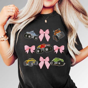 Lady Wearing a Black Comfort Colors t-shirt featuring monster trucks in various colors, including blue, orange, red, yellow, and green. Pink bows are interspersed between the truck in three rows. The shirt has a crew neck and short sleeves.