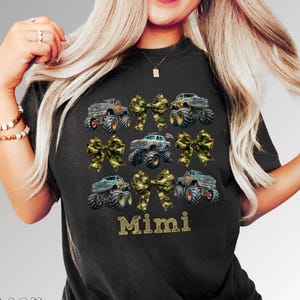 Woman wearing a black t-shirt featuring a design of monster trucks and camouflage bows. The word "Mimi" is printed in gold lettering below the trucks. The shirt is a comfortable, casual style.