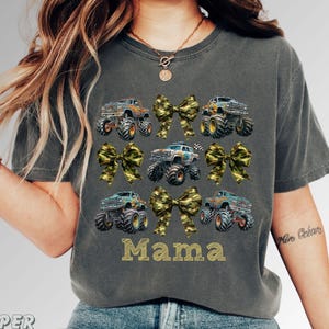 Pictured is a woman wearing a dark gray t-shirt featuring a design of monster trucks and camouflage bows. The word "Mama" is printed in gold block letters. The shirt has a crew neck and short sleeves.