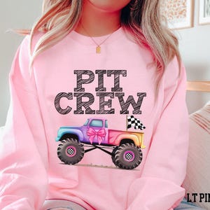 model is wearing a pretty pink monster truck sweatshirt with watercolor rainbow colors and coquette  black and white checkered flag sketch big graphic letters family matching different version for male and female pink for the girls and purple/blue for the guys
