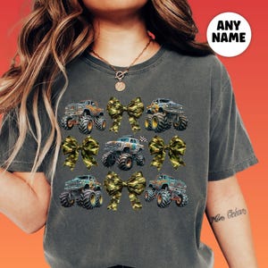 woman wearing a comfort colors pepper vintage shirt with camouflage camo coquette bows and monster trucks in a collage style grid. Perfect for Monster truck show race day, birthday party, gift for Monster truck lover. A girly Tough Girl Power Look and feel. Mommy and Me matching set with personalized optional custom name. Monster truck Girls Night Out group shirt idea, Boy Mama shirt.