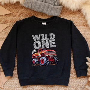 Op de afbeelding: Zwart sweatshirt met de witte tekst "WILD ONE". Een rode monstertruck met blauwe banden en een geblokte vlag is eronder gedrukt. De sweatshirt heeft lange mouwen en een ronde hals.