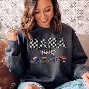 Op de afbeelding: Een donkergrijs sweatshirt met het woord "MAMA" in zwart-wit geruite letters. Onder het woord "MAMA" staan vier monstertrucks met geruite vlaggen.