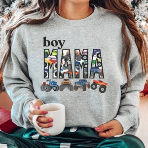 Op de afbeelding: Grijs sweatshirt met de tekst "boy mama" in zwarte letters. De letters zijn gevuld met een patroon van kleurrijke monstertrucks.