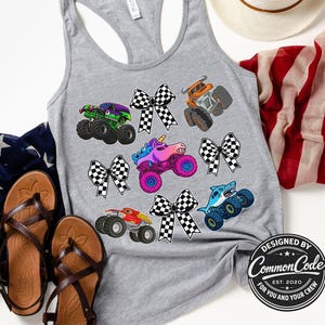 May include: A gray tank top featuring colorful monster truck graphics, including a green, purple, orange, red, and blue truck design. The shirt also has black and white checkered bows. A pair of brown sandals and a red, white, and blue scarf are in the background.