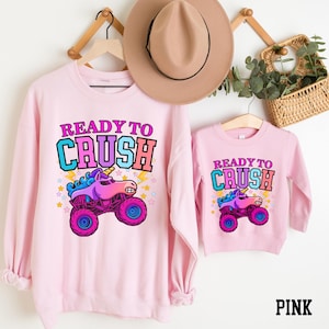 Pu&ograve; includere: Felpe rosa con la scritta "READY TO CRUSH" e una grafica di camion mostro unicorno dei cartoni animati. Una &egrave; per adulti, l'altra per bambini. Un cappello marrone e un cesto di verde sono sullo sfondo.