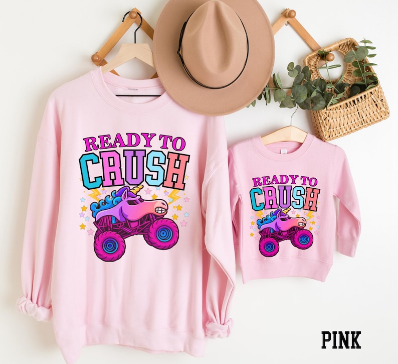 Pu&ograve; includere: Felpe rosa con la scritta "READY TO CRUSH" e una grafica di camion mostro unicorno dei cartoni animati. Una &egrave; per adulti, l'altra per bambini. Un cappello marrone e un cesto di verde sono sullo sfondo.