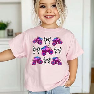 May include: A light pink t-shirt featuring a repeating pattern of monster trucks with unicorn horns, checkered bows, and vibrant colors. The monster trucks have pink, purple, and blue accents.