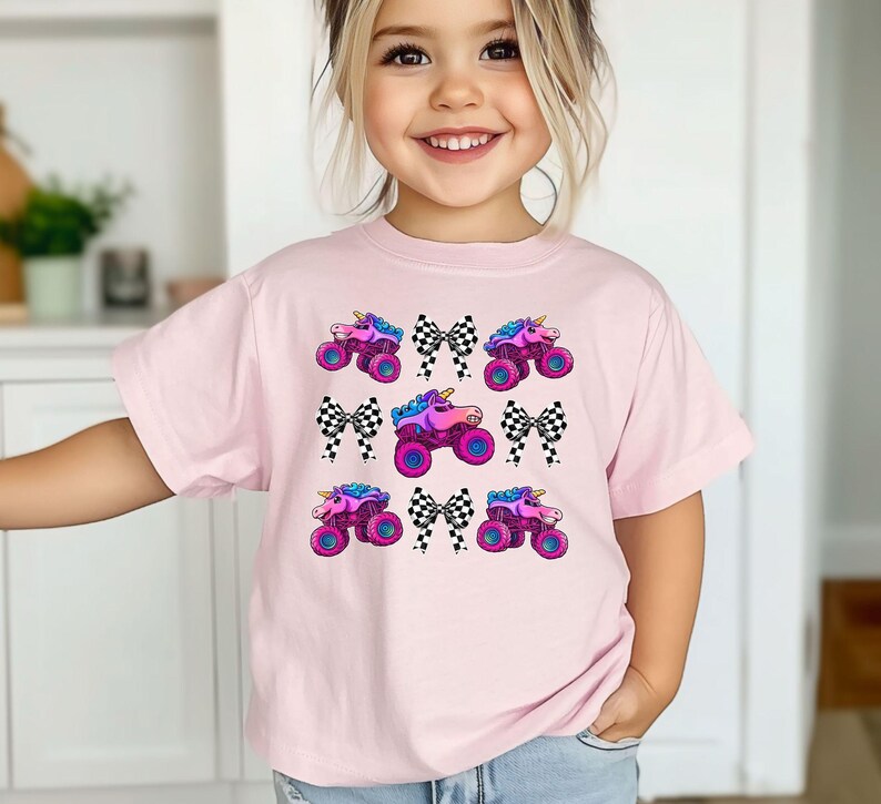 May include: A light pink t-shirt featuring a repeating pattern of monster trucks with unicorn horns, checkered bows, and vibrant colors. The monster trucks have pink, purple, and blue accents.
